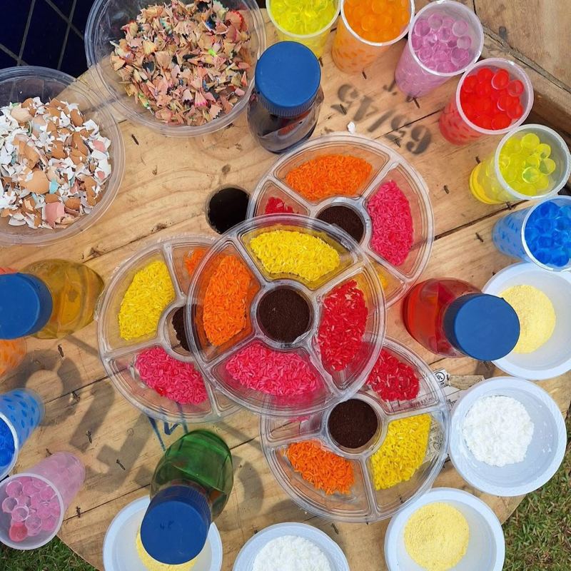Mesa de exploração com arroz colorido, bolinhas de gel e materiais sensoriais montada por Edineia
