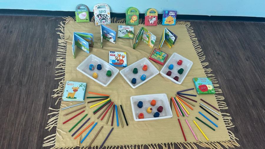 Espaço com livros de bebê, potes com tampinhas e lápis de cor sobre tapete
